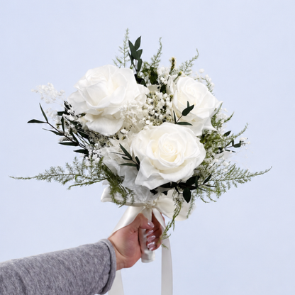 Hand holding the Opal Promise Petit Preserved Flower Bouquet showing 3 white roses with baby's breath and asparagus fern against a soft blue background