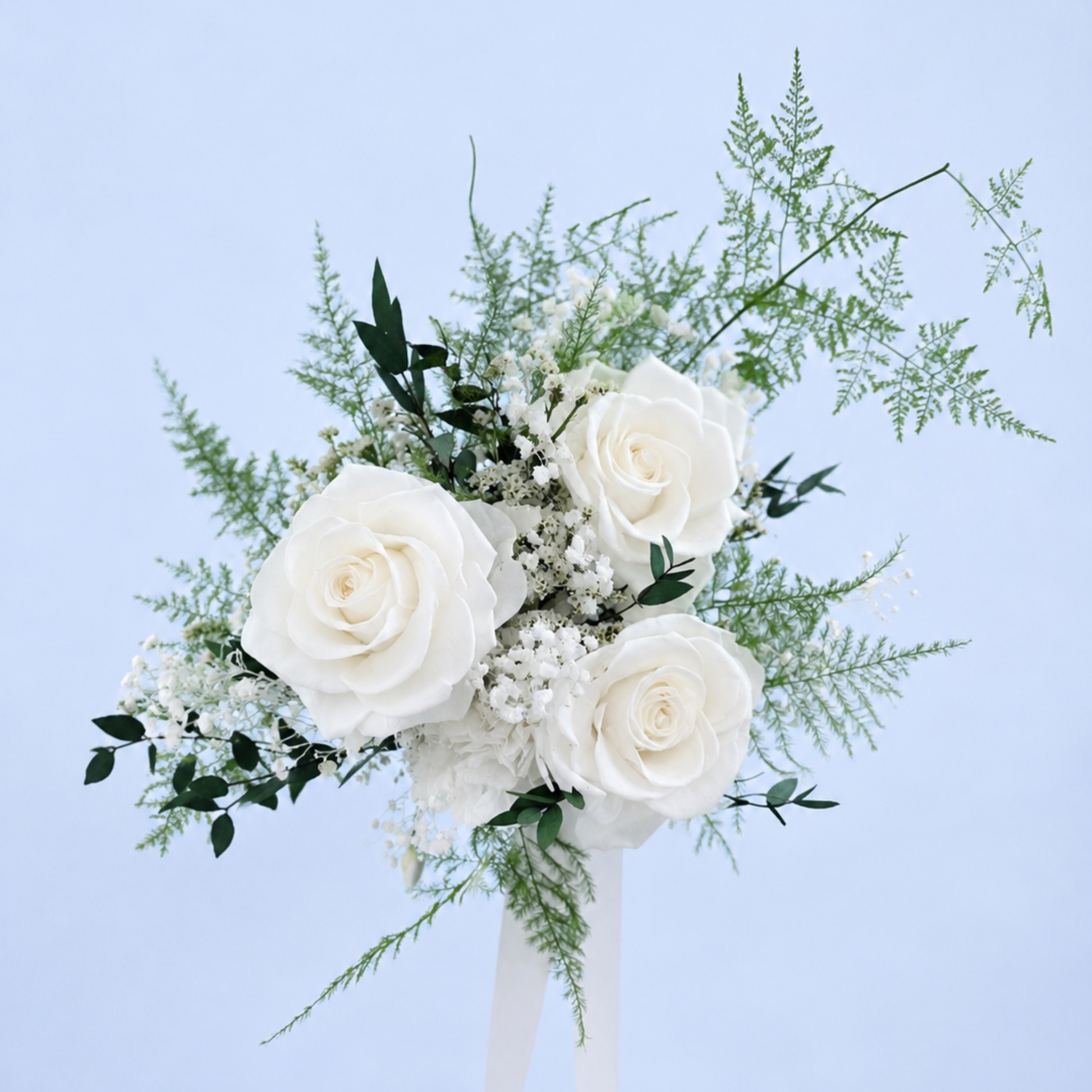 Aerial view of Opal Promise Petit Preserved Flower Bouquet featuring 3 white roses, baby's breath and trailing asparagus fern against a soft blue background