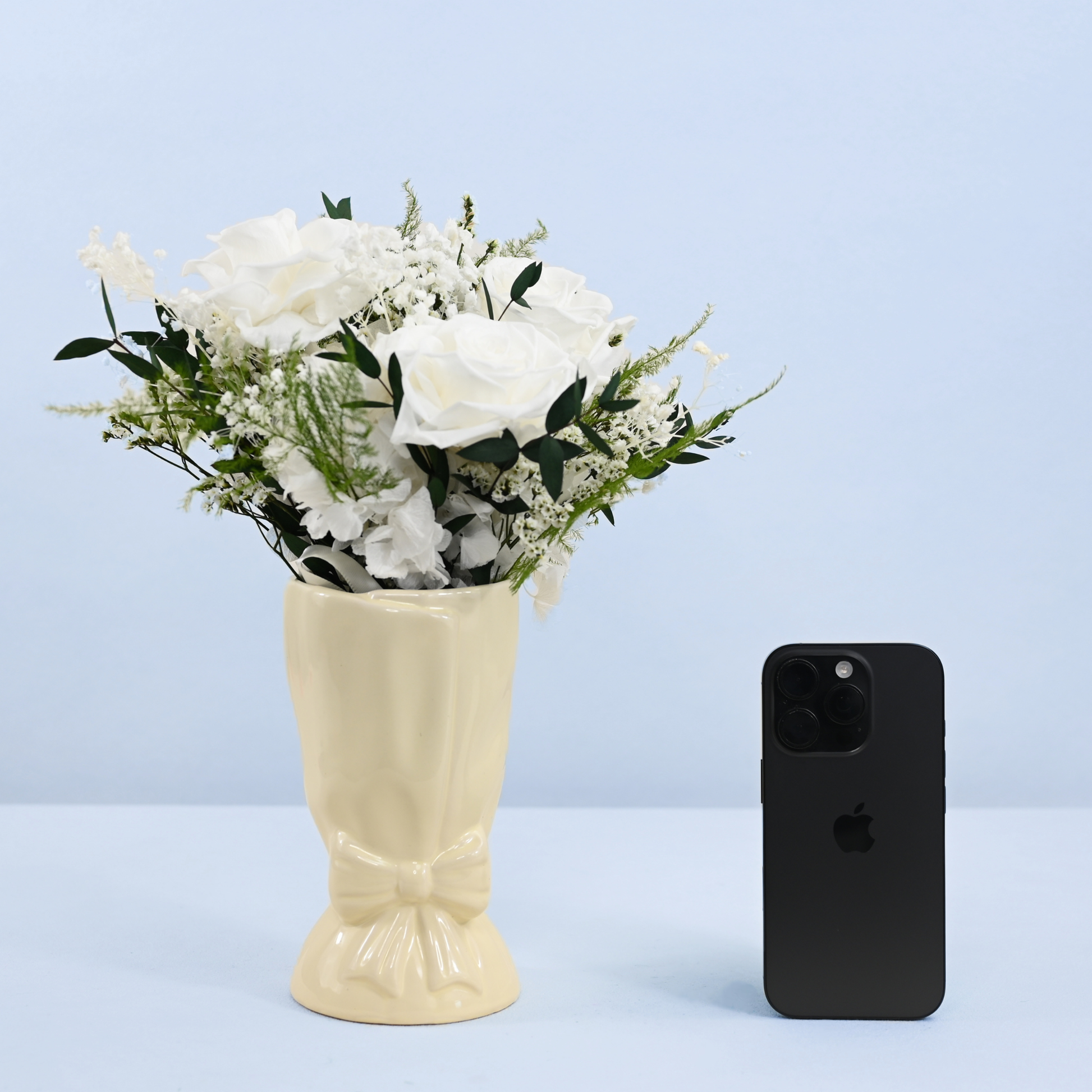 Opal Promise Petit Preserved Flower Bouquet in cream ceramic bow vase shown next to an iPhone for size reference
