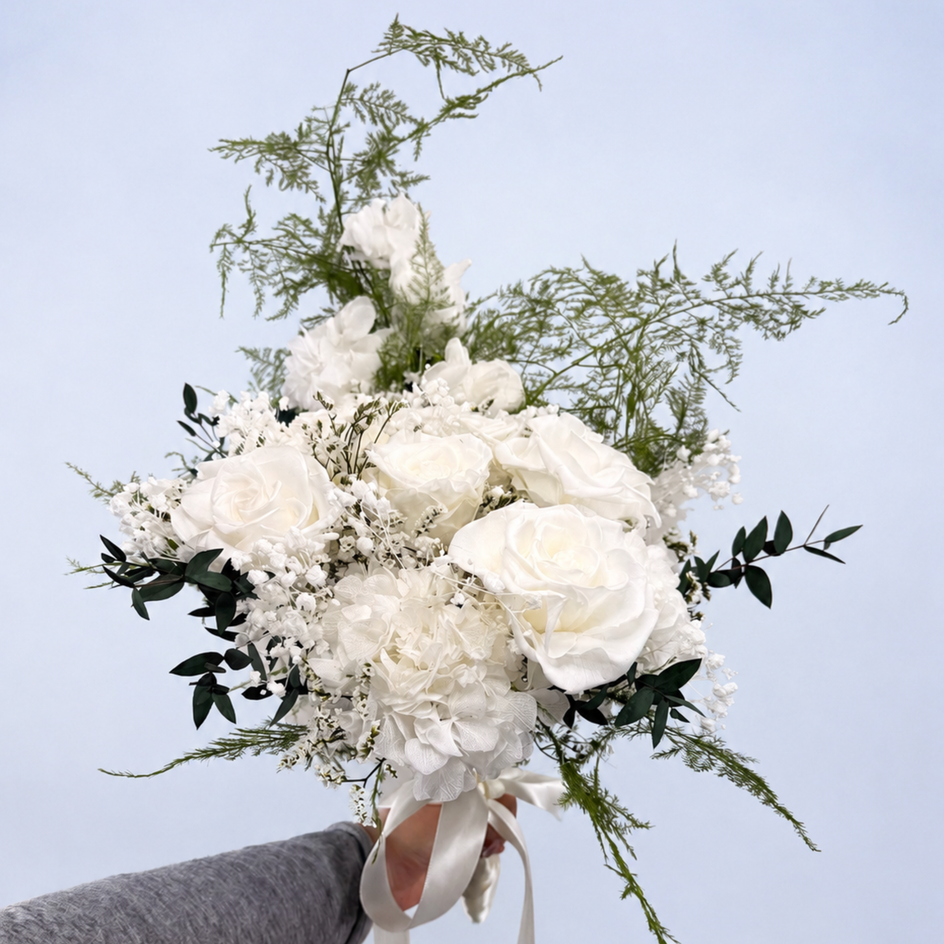 Opal Promise Preserved Flower Bouquet held up by hand showing white roses, hydrangea, baby's breath and trailing asparagus fern against a soft blue background
