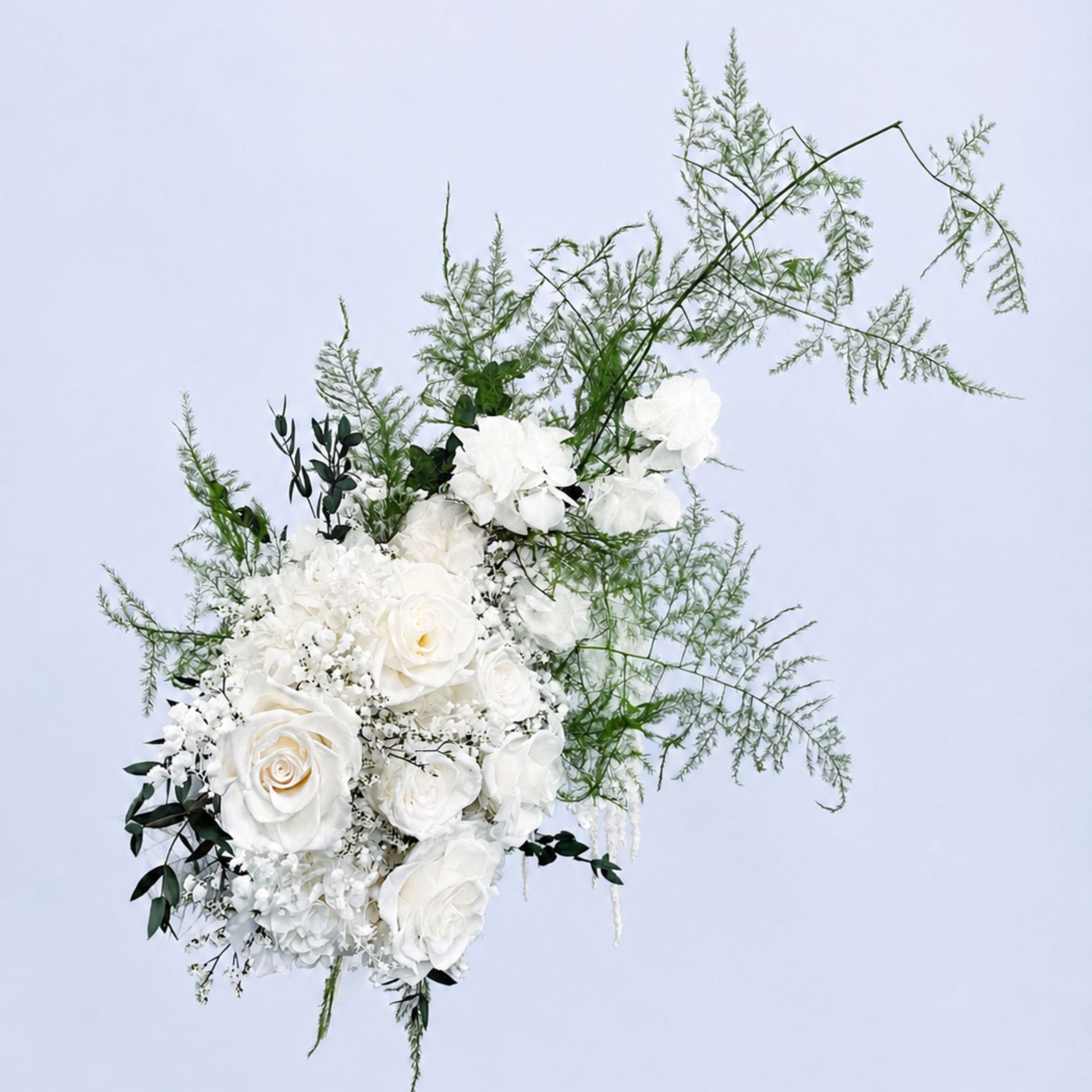 Aerial view of Opal Promise Preserved Flower Bouquet featuring white roses, hydrangea, baby's breath and flowing asparagus fern against a soft blue background