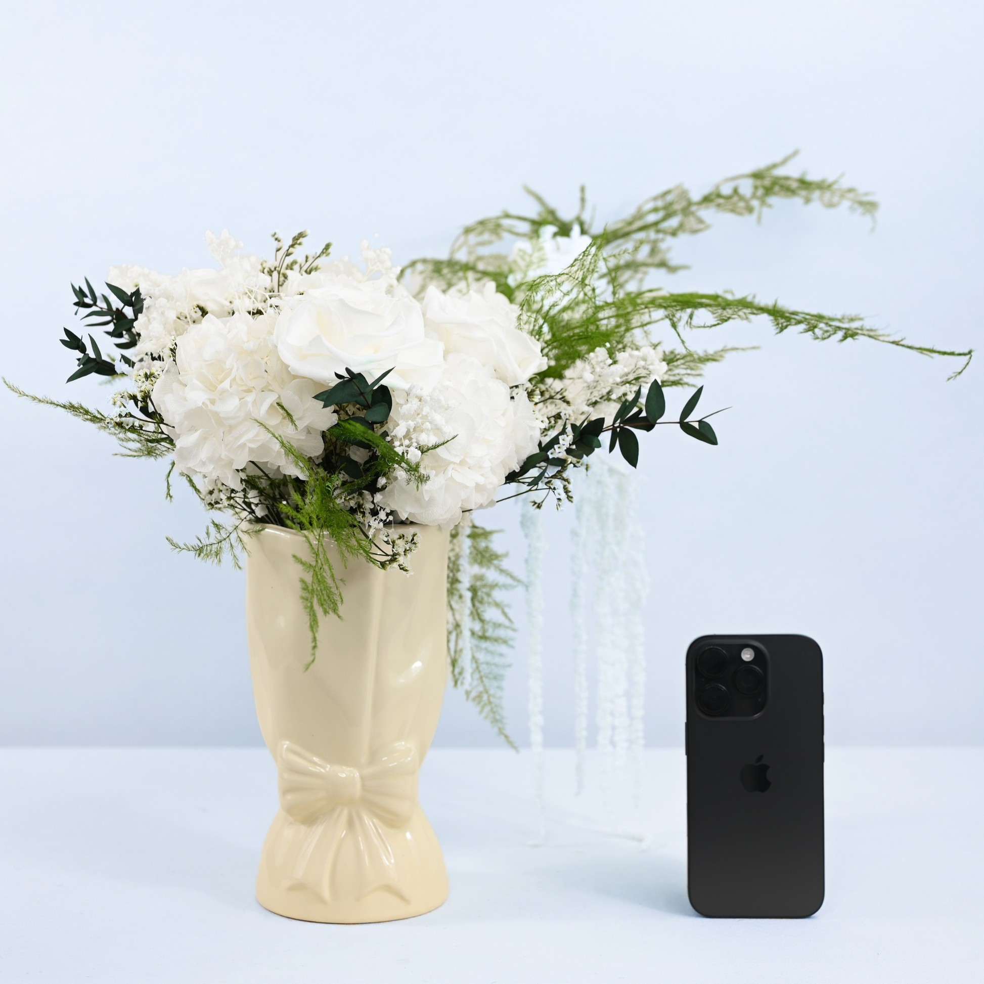 Opal Promise Preserved Flower Bouquet in cream ceramic bow vase shown next to an iPhone for size reference