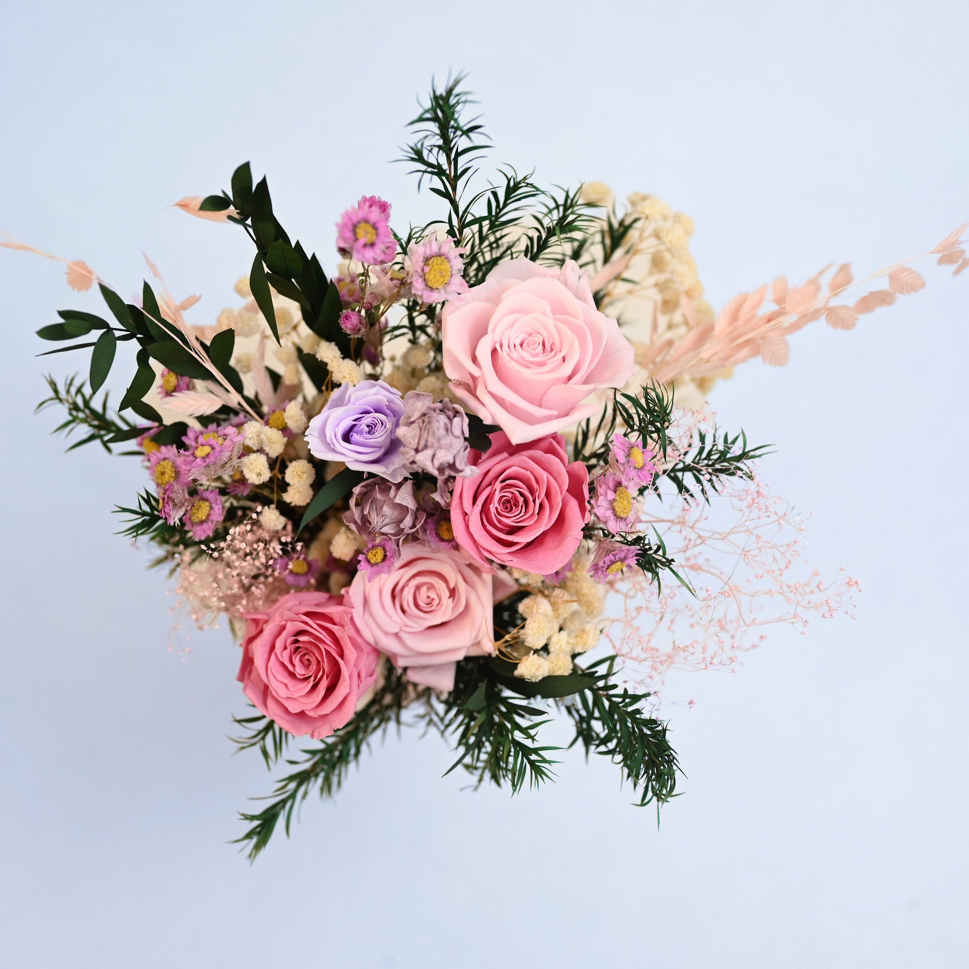 A preserved floral arrangement featuring pink roses, lavender lisianthus, cream baby's breath, and green foliage, photographed from directly above against a solid light blue background.