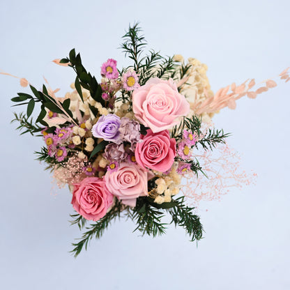 A preserved floral arrangement featuring pink roses, lavender lisianthus, cream baby's breath, and green foliage, photographed from directly above against a solid light blue background.
