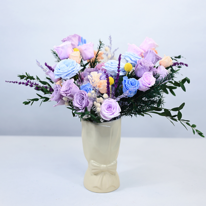 Lavender Sky Preserved Flower Bouquet with blue and lavender roses, hydrangea, and craspedia displayed in a cream ceramic bow vase