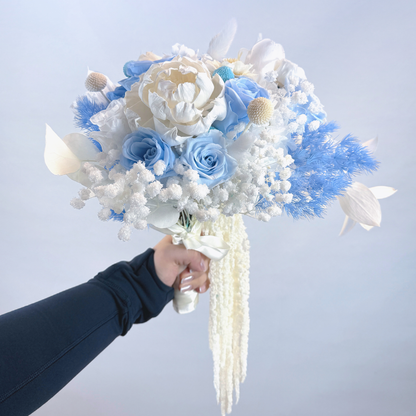 Hand holding Sapphire Veil Preserved Flower Bouquet featuring blue roses, white pith flower, eryngium and cascading white amaranthus against a soft blue background