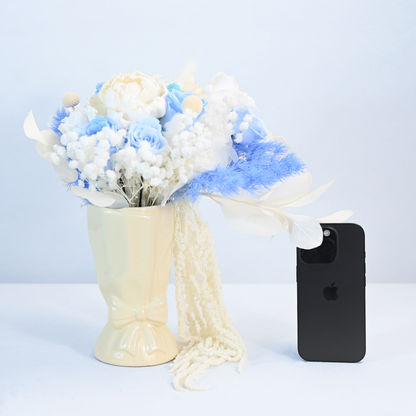 Sapphire Veil Preserved Flower Bouquet laid beside a cream ceramic bow vase with cascading white amaranthus and iPhone for size reference