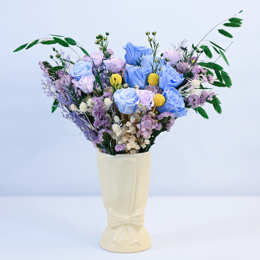 Moonstone Garden Preserved Flower Bouquet with blue and lavender roses, craspedia, and statice in a cream ceramic bow vase