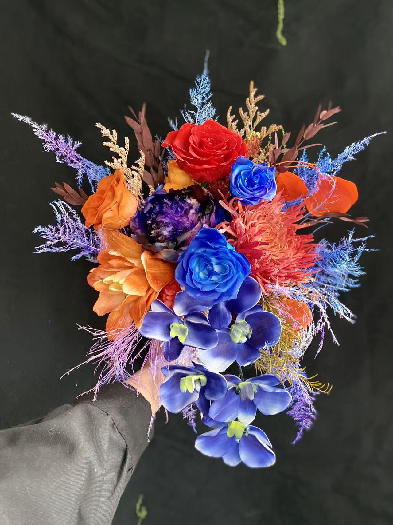 Tourmaline Cascade Preserved Flower Bouquet hand-held arrangement featuring cascading blue orchids, red and blue roses, orange lily, purple peony, and jewel-tone fern fillers