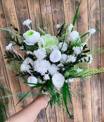 Laurel Muse Preserved Flower Bouquet hand-held arrangement of white roses, carnations, hydrangea, and statice with lush green fan palm and asparagus fern against a wooden background