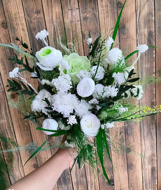 Laurel Muse Preserved Flower Bouquet hand-held arrangement of white roses, carnations, hydrangea, and statice with lush green fan palm and asparagus fern against a wooden background
