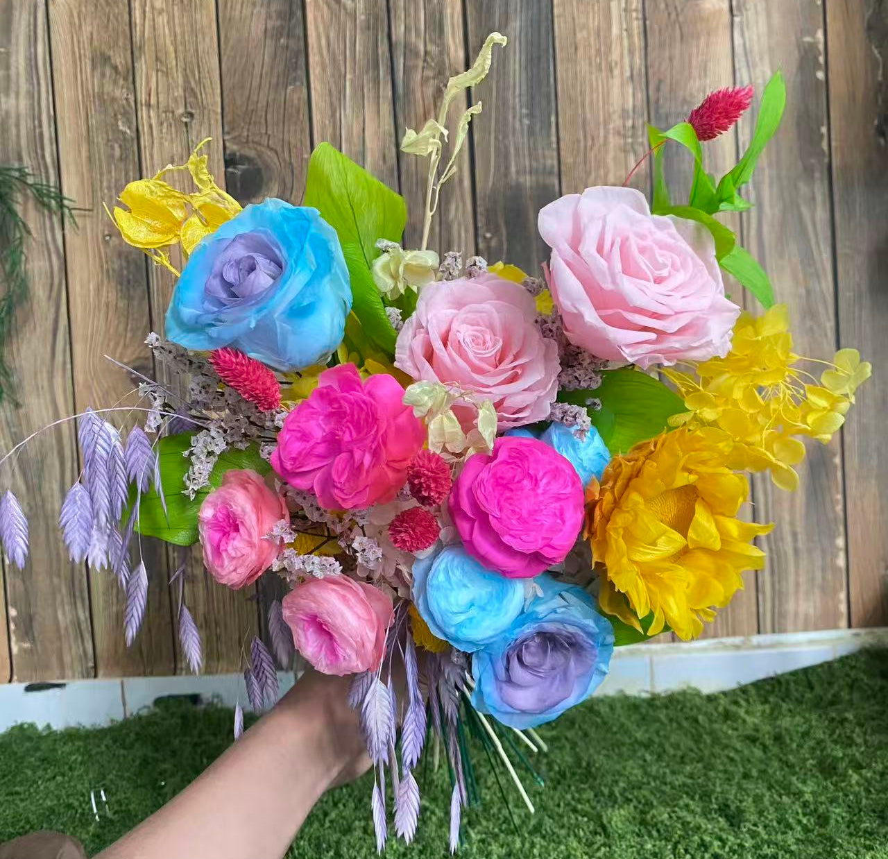 Carnival Bloom Preserved Flower Bouquet hand-held arrangement of pink and blue roses, hot pink and blue Austin roses, yellow sunflower, and pink and yellow hydrangea with green leaves and colorful fillers