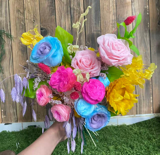 Carnival Bloom Preserved Flower Bouquet hand-held arrangement of pink and blue roses, hot pink and blue Austin roses, yellow sunflower, and pink and yellow hydrangea with green leaves and colorful fillers