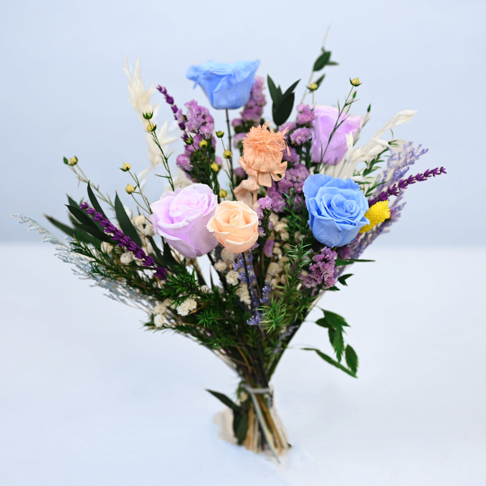 A pastel-colored artificial flower bouquet featuring blue, pink, and peach roses with purple filler flowers and green foliage, tied with a simple beige ribbon and set against a clean white background.