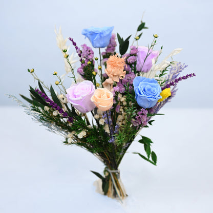 A pastel-colored artificial flower bouquet featuring blue, pink, and peach roses with purple filler flowers and green foliage, tied with a simple beige ribbon and set against a clean white background.