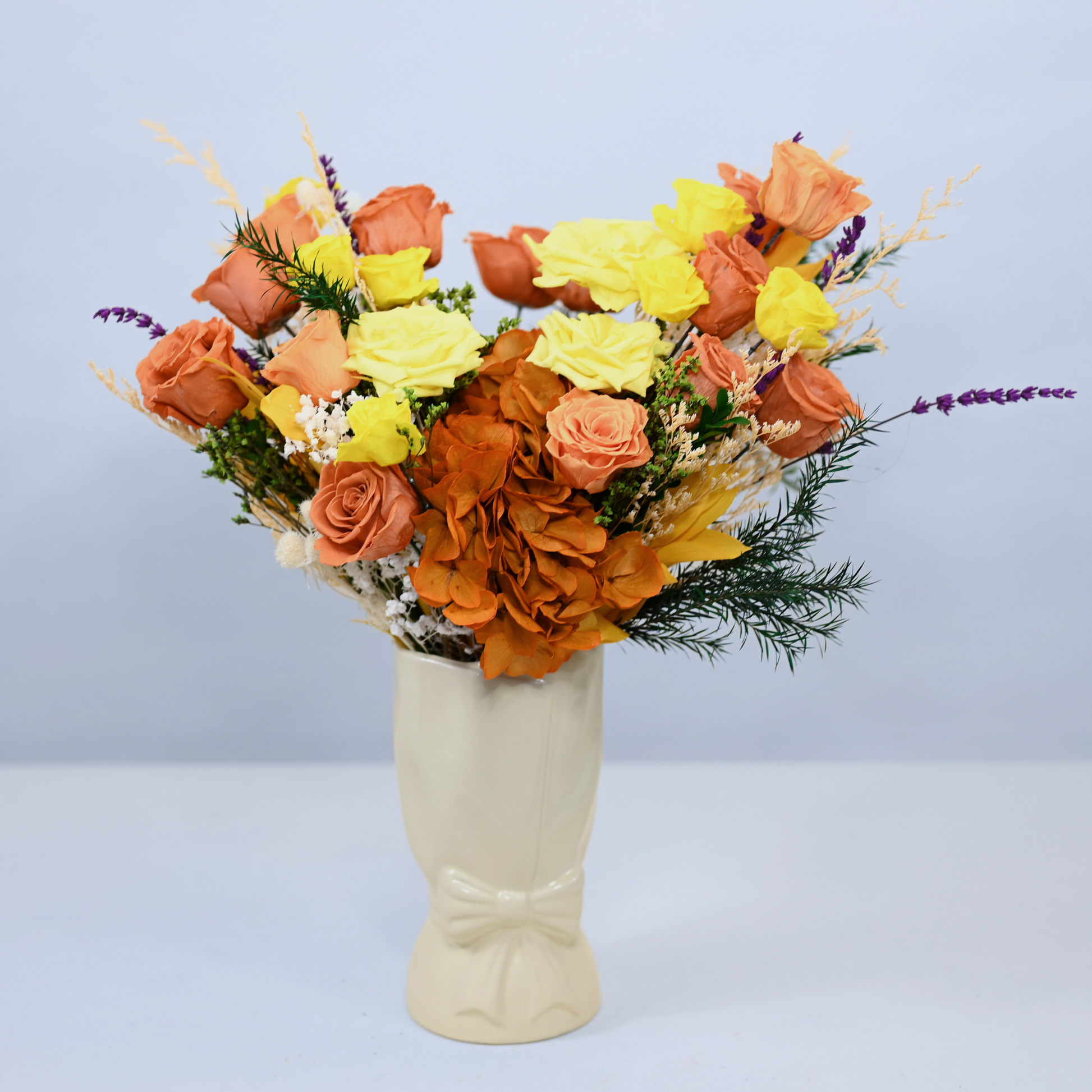 Golden Hour Preserved Flower Bouquet with orange and yellow roses, orange hydrangea and purple lavender displayed in a cream ceramic bow vase, front view