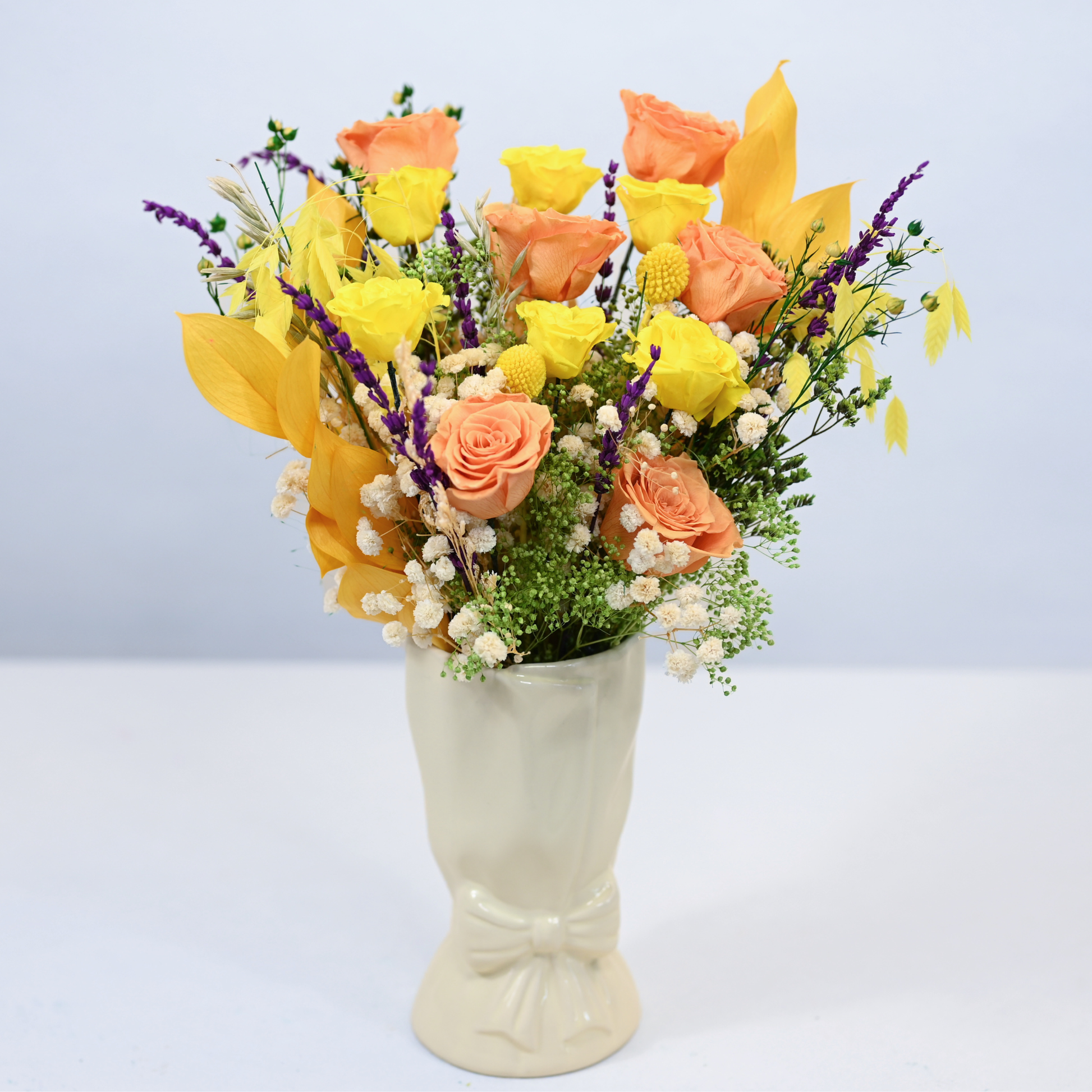 Citrus Meadow Preserved Flower Bouquet with orange and yellow roses, craspedia and purple lavender in a cream ceramic bow vase, front view