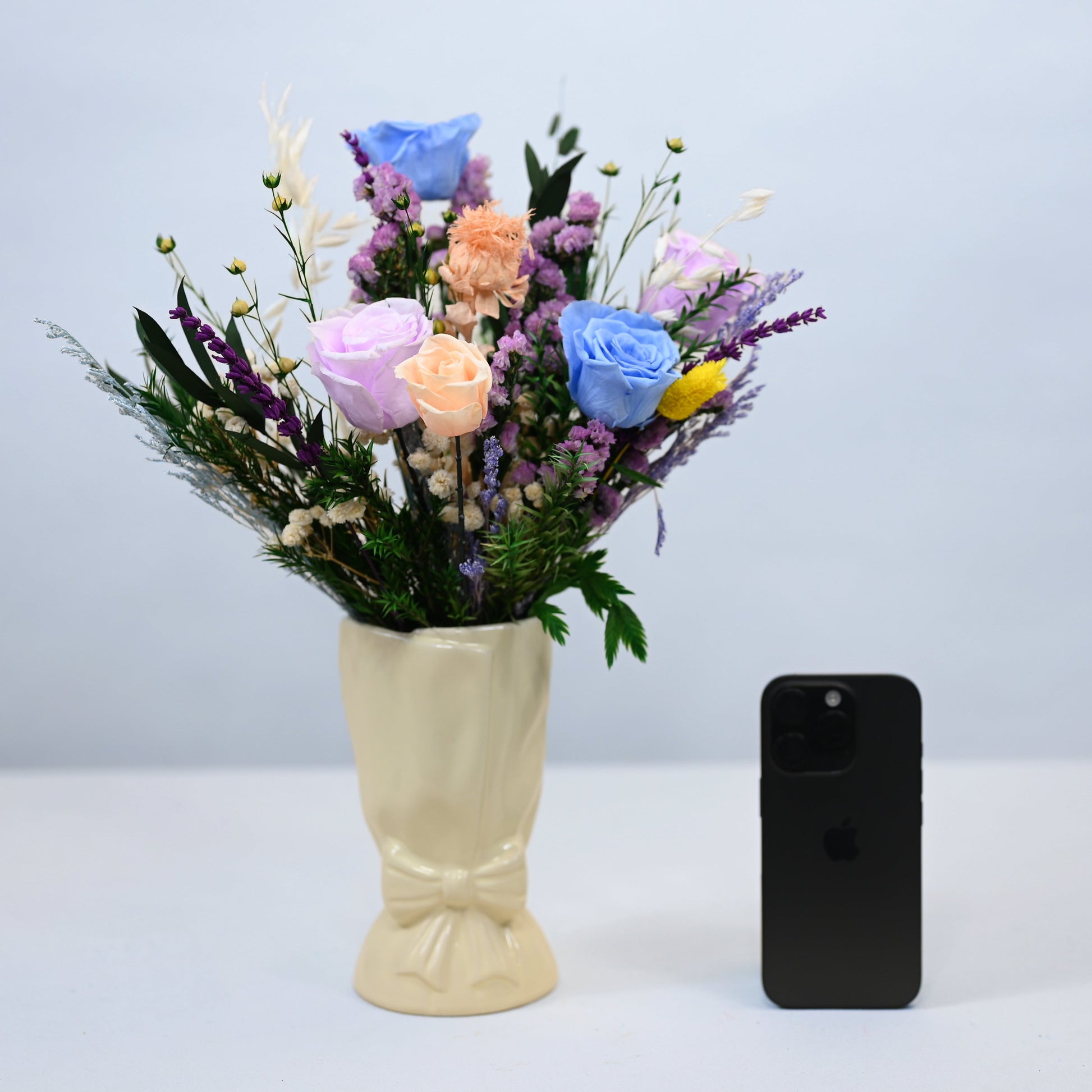 A pastel flower bouquet in a cream-colored vase, displayed next to a black smartphone on a light gray surface against a matching gray background.