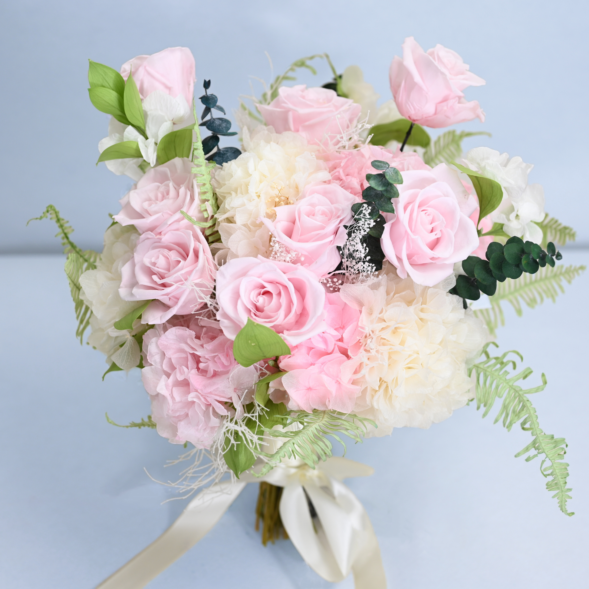 Rose Villa Preserved Flower Bouquet close-up showing pink roses, carnations and leatherleaf fern with cream satin ribbon against a soft blue background