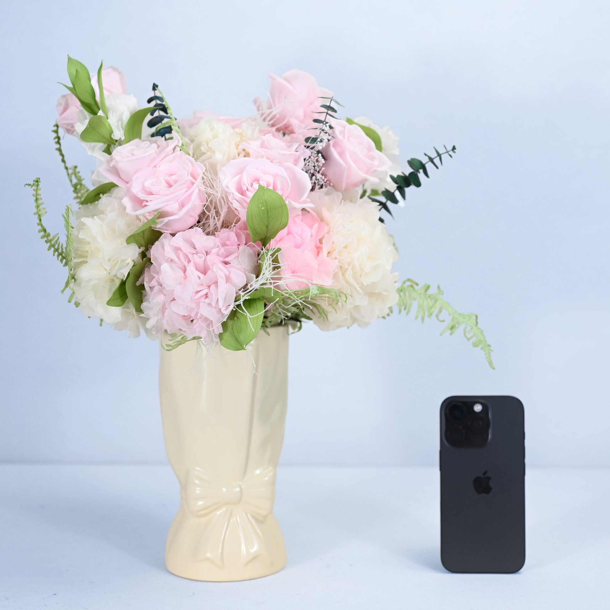 Rose Villa Preserved Flower Bouquet in cream ceramic bow vase shown next to an iPhone for size reference