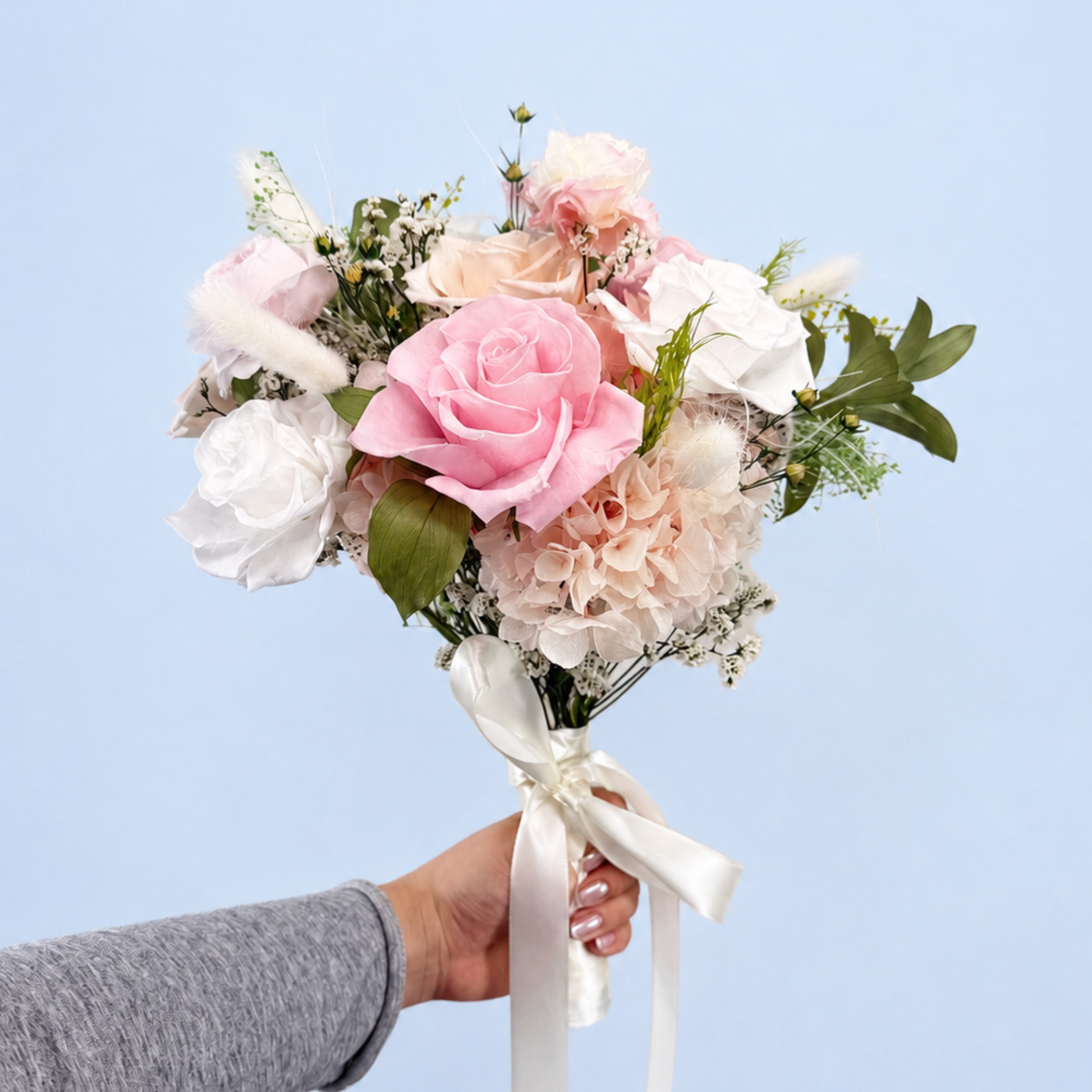 Hand holding Rose Villa Petit Preserved Flower Bouquet showing pink, white and peach roses with hydrangea and pampas grass against a soft blue background