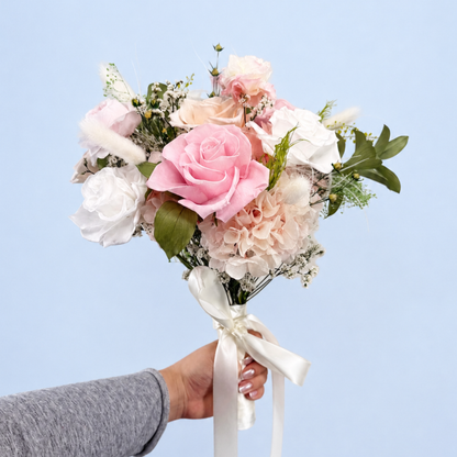 Hand holding Rose Villa Petit Preserved Flower Bouquet showing pink, white and peach roses with hydrangea and pampas grass against a soft blue background