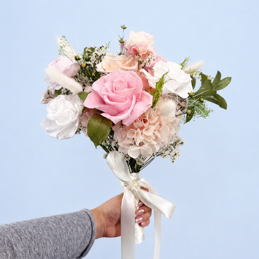 Hand holding Rose Villa Petit Preserved Flower Bouquet showing pink, white and peach roses with hydrangea and pampas grass against a soft blue background