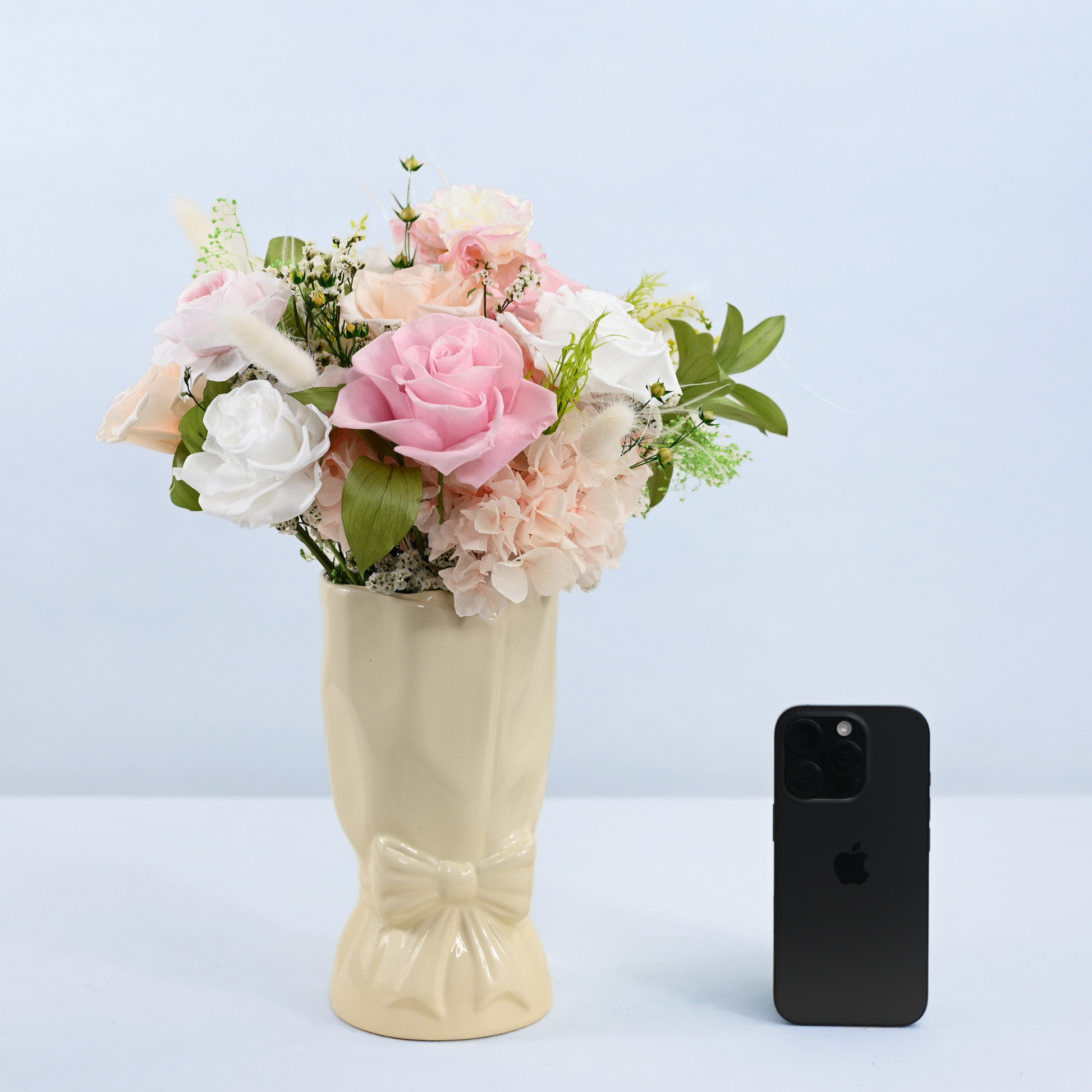 Rose Villa Petit Preserved Flower Bouquet in cream ceramic bow vase shown next to an iPhone for size reference