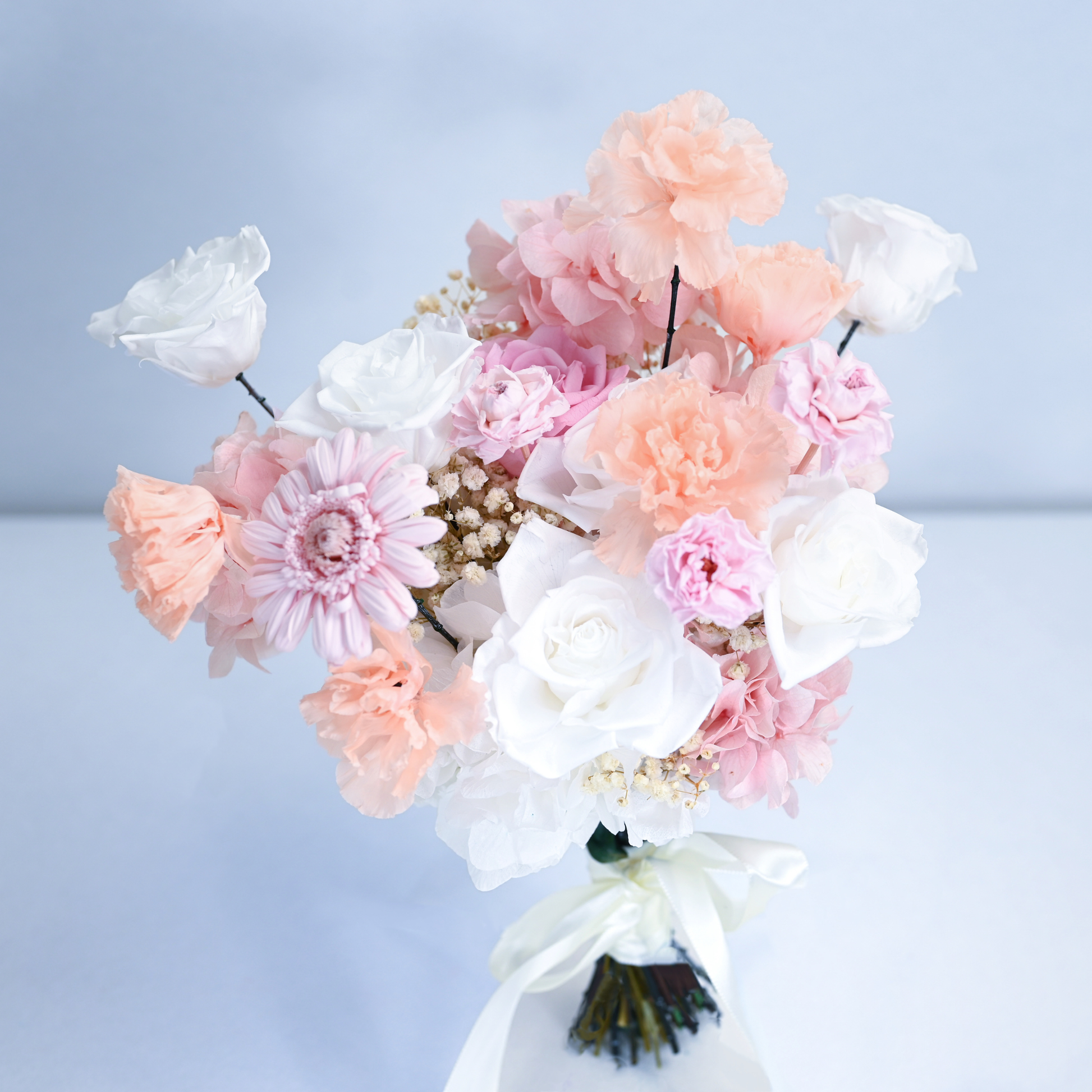 Blush Reverie Preserved Flower Bouquet showing white and pink roses, peach lisianthus, gerbera daisy and hydrangea with cream satin ribbon against a soft blue background