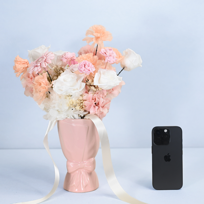 Blush Reverie Preserved Flower Bouquet in a pink ceramic bow vase shown next to an iPhone for size reference