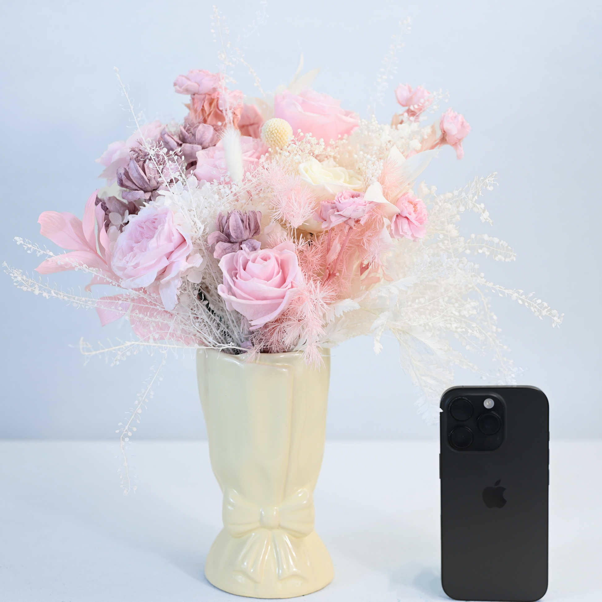 Blush Muse Preserved Flower Bouquet in cream ceramic bow vase shown next to an iPhone for size reference