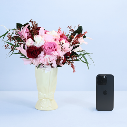Crimson Blush Preserved Flower Bouquet in cream ceramic bow vase shown next to an iPhone for size reference