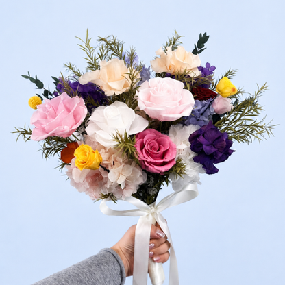 Hand holding Rainbow Garden Preserved Flower Bouquet showing multicolor roses, purple hydrangea, craspedia and asparagus fern against a soft blue background
