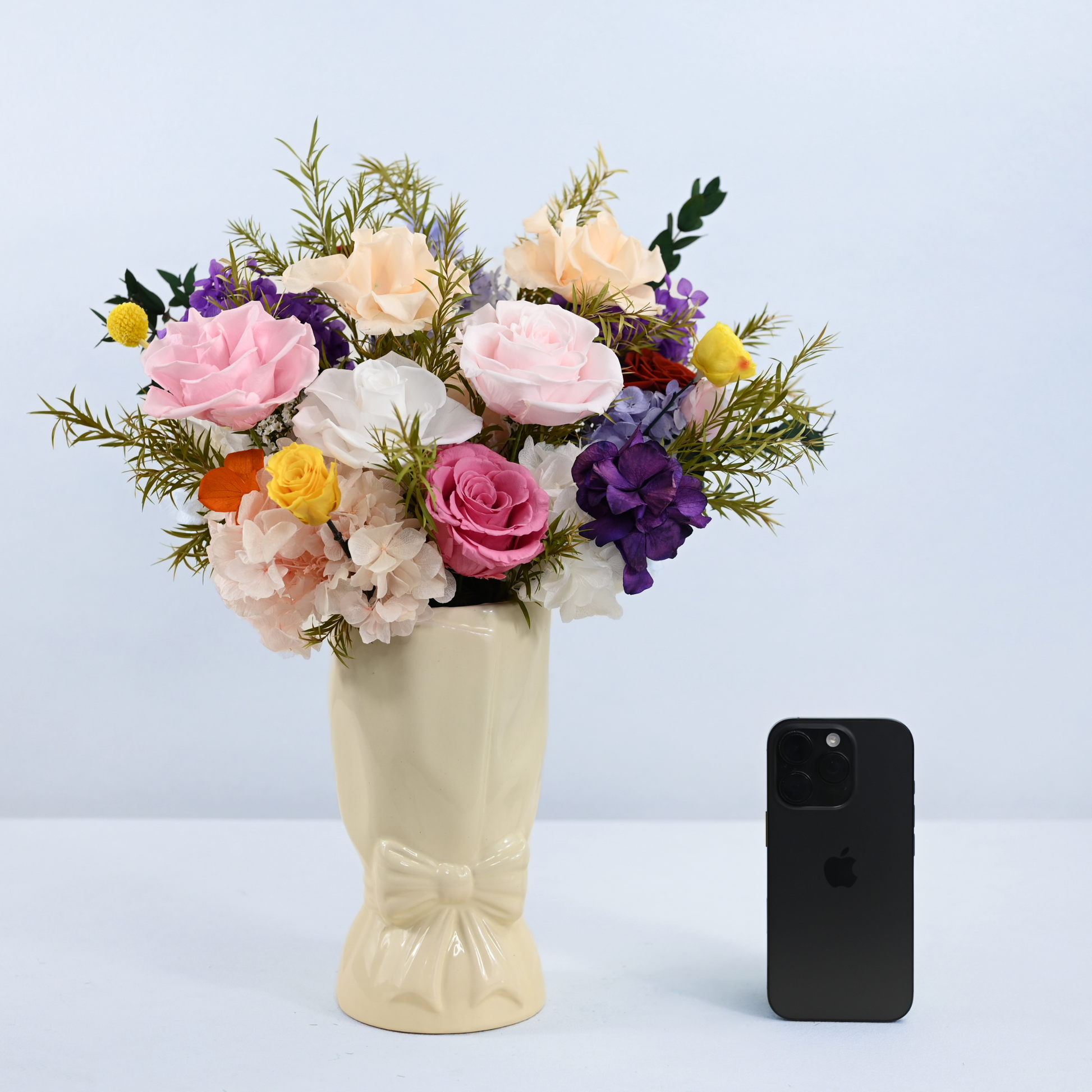 Rainbow Garden Preserved Flower Bouquet in cream ceramic bow vase shown next to an iPhone for size reference
