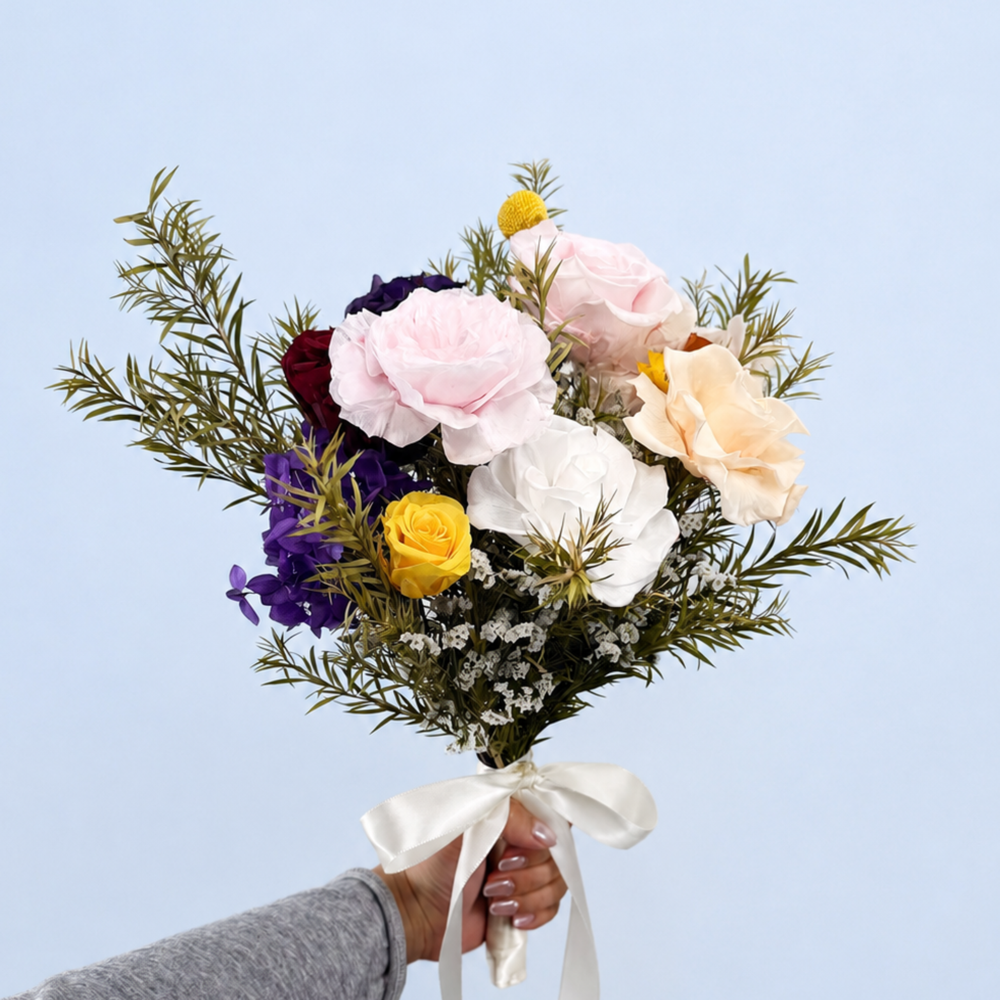 Hand holding Rainbow Garden Petit Preserved Flower Bouquet showing multicolor roses, purple hydrangea, craspedia and asparagus fern against a soft blue background
