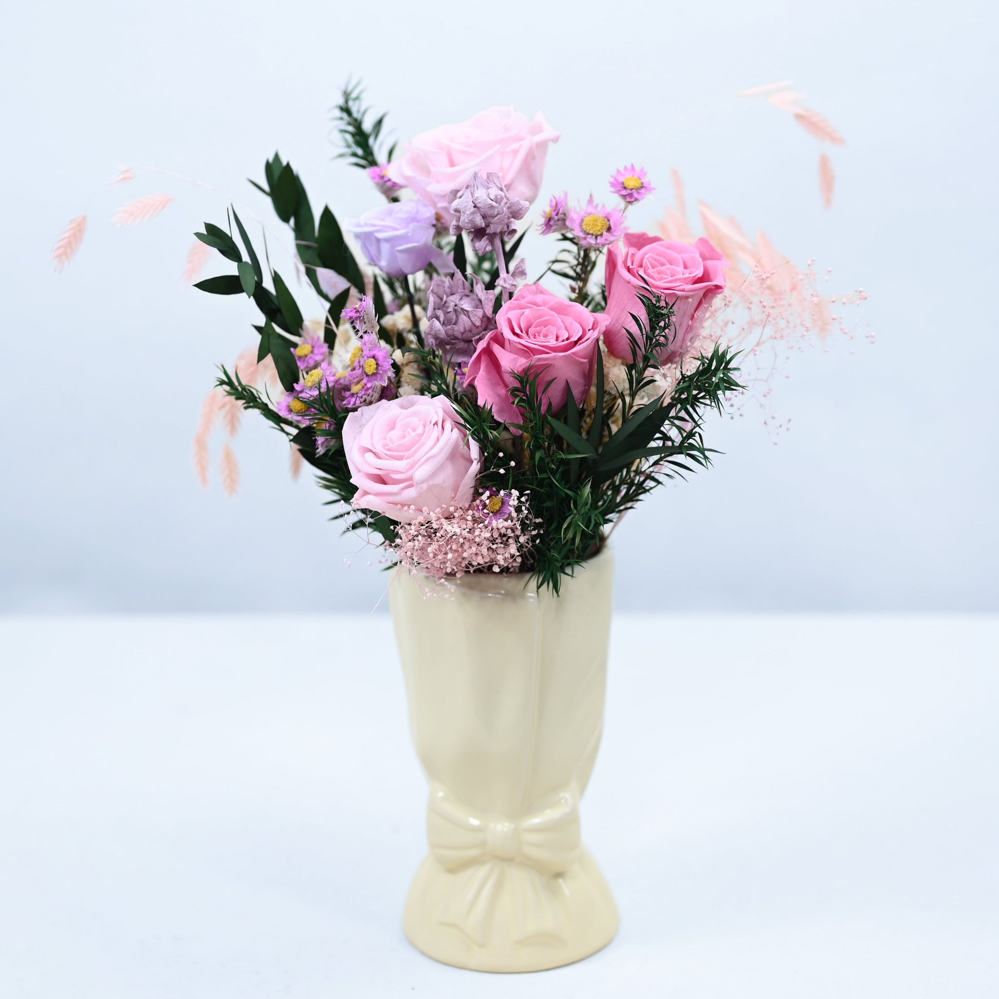 A pastel pink and lavender floral arrangement with green foliage, displayed in a cream-colored ceramic vase against a light blue-gray background.
