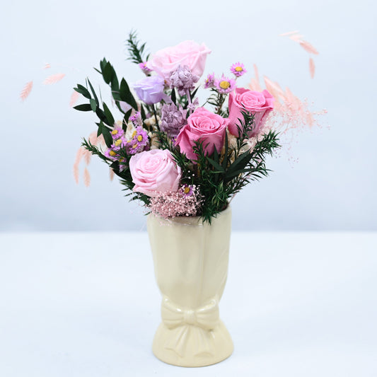 A pastel pink and lavender floral arrangement with green foliage, displayed in a cream-colored ceramic vase against a light blue-gray background.